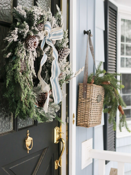 A wreath hangs on a front door and a basket of evergreen branches is hung next to it