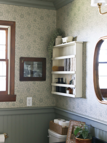 A white shelf hangs on a bathroom wall above the toilet and next to a framed mirror