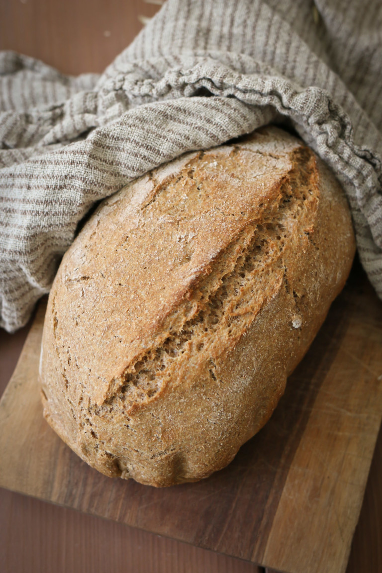 A loaf of rustic artisan bread made with freshly milled flour rests next to a linen tea towel