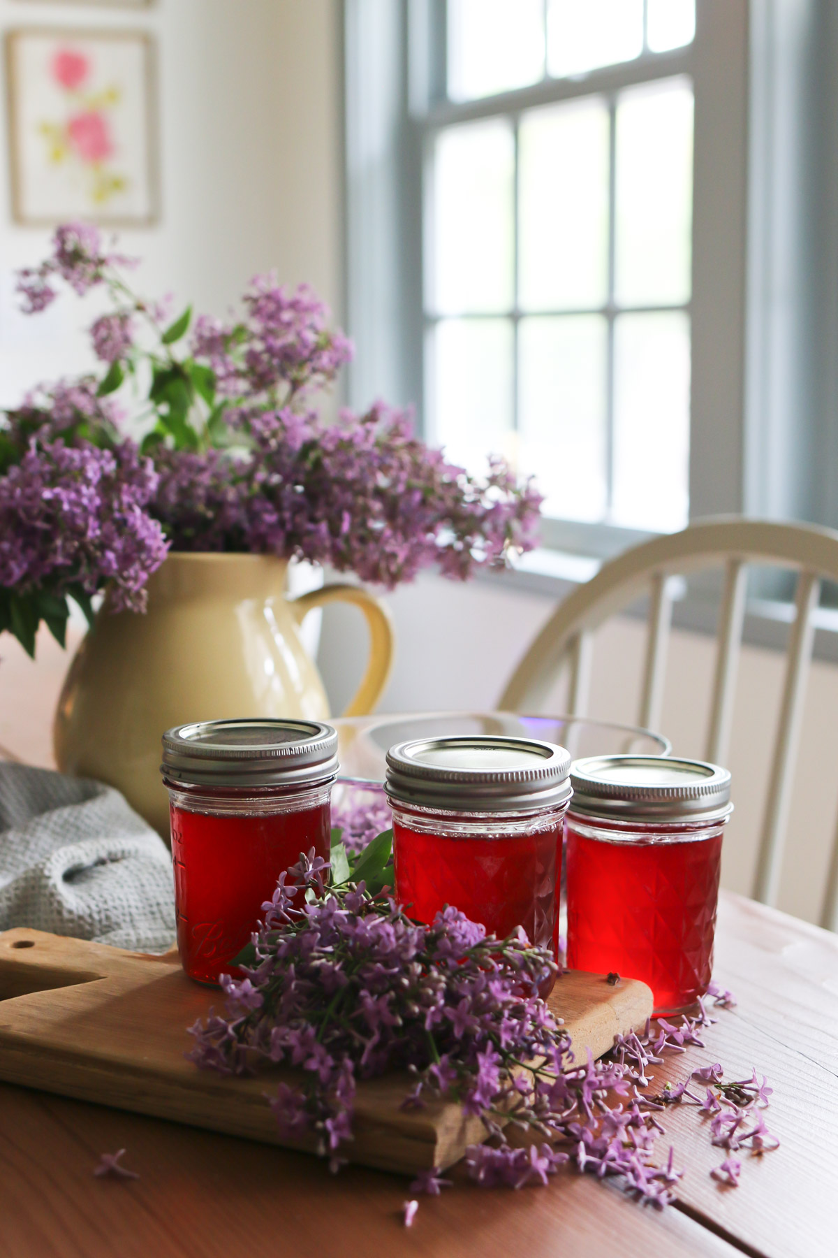 A kitchen table holds a vase of lilac flowers along with 3 jars of a lilac jelly recipe