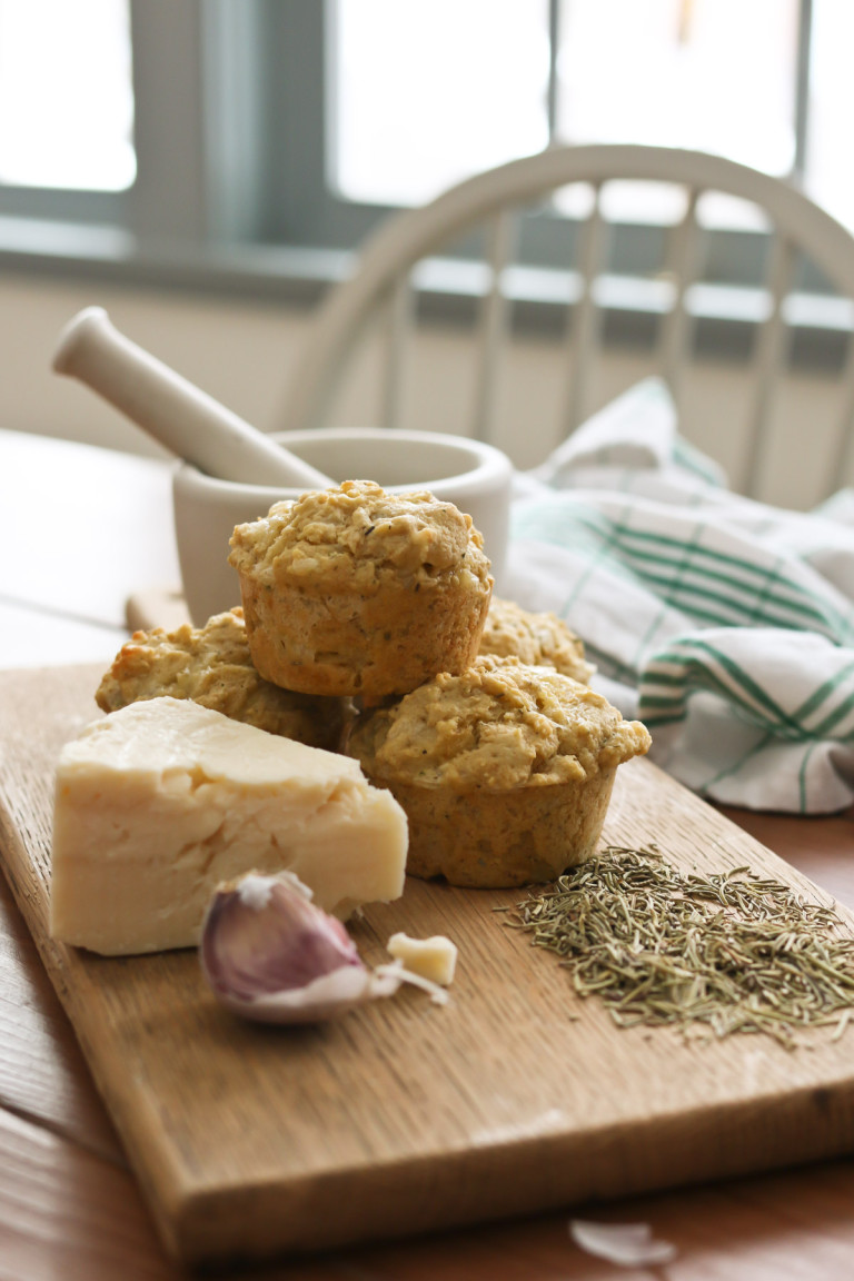 Savory herb and cheese muffins are arranged next to a cheese wedge and a pile of herbs